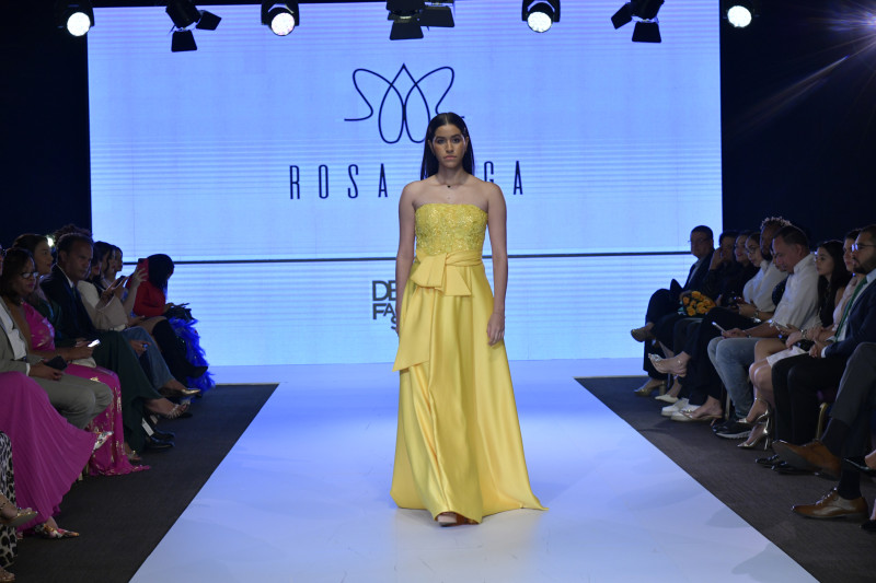 Pasarela oficial | DBA Fashion Show – Rosa Boga Modelo desfila con vestido amarillo en pasarela del DBA Fashion Show, con logo de Rosa Boga al fondo.